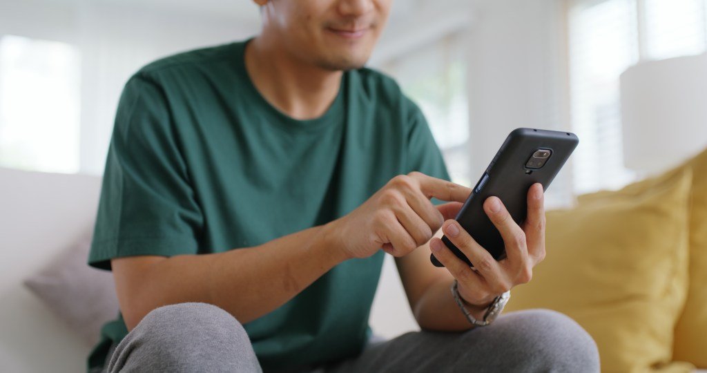 An adult Asian man sitting on the sofa at home, smiling and swiping right on a dating app on his phone