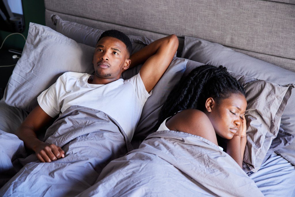 Young African man looking nervous and unhappy, lying on the bed next to his wife sleeping peacefully in the morning