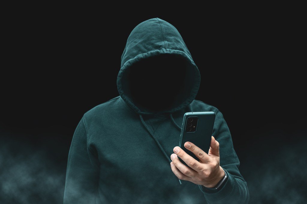 The outline of a faceless, hooded character, probably a hacker or cybercriminal, holding a smartphone