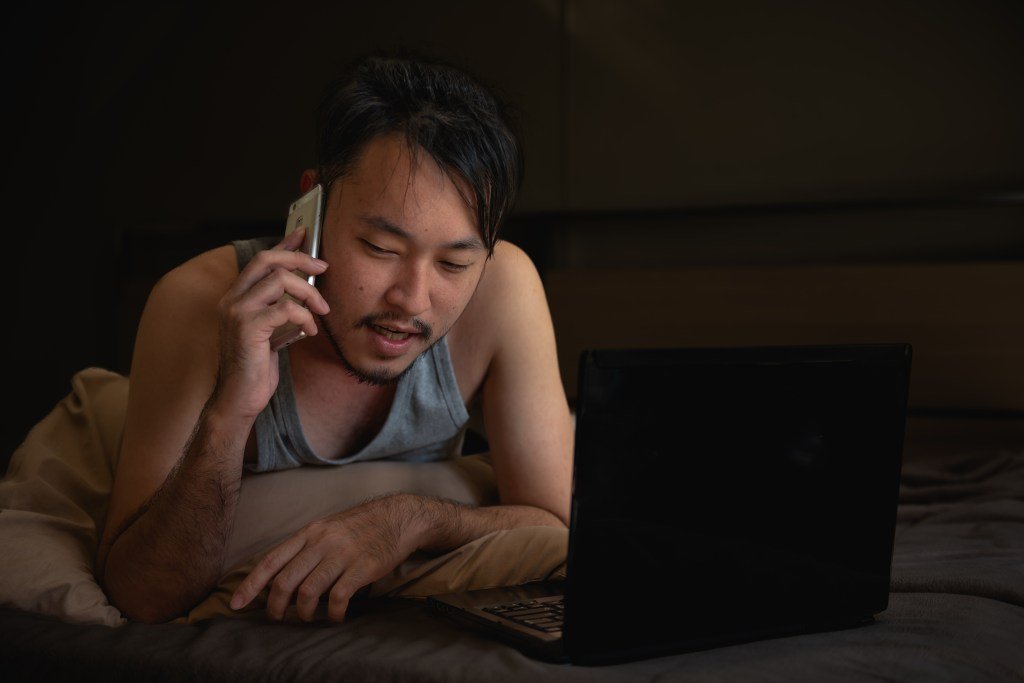 Young Asian man using laptop in bed at night reflecting the loneliness of young American man