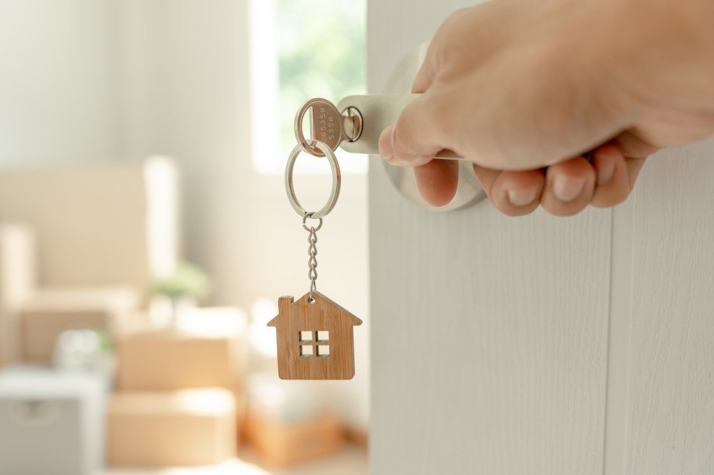 One hand unlocks the door with a house-shaped keychain.