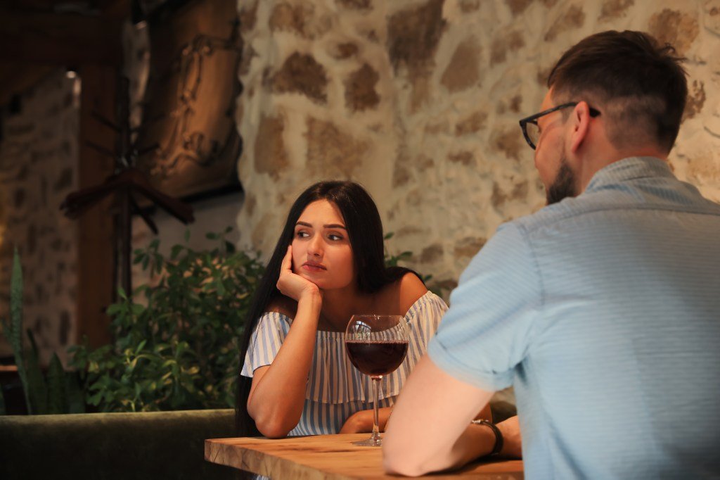 Young woman with long dark hair in striped off-shoulder top looks bored on a man's date in a cafe.